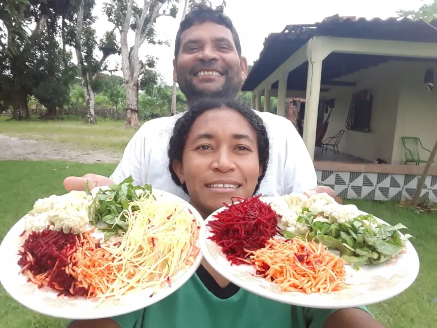 Maria do Rosário e o esposo em casa, no sítio, segurando pratos coloridos de comida da terra